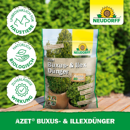 Um saco de Neudorff Shop Azet Buxus- & IlexDünger, um fertilizante orgânico para buxos e azevinhos, está ao ar livre sobre uma mesa. A embalagem indica a segurança para animais de estimação e ingredientes naturais, enquanto ao fundo se veem arbustos verdes exuberantes.