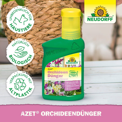 Uma garrafa verde Azet OrchideenDünger líquido da Neudorff Shop está sobre uma superfície rosa. Seu rótulo de plástico reciclado, 100% biológico e seguro para animais promete flores esplêndidas. Ao fundo, vê-se uma cesta trançada e uma planta.