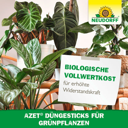 No interior, várias plantas verdes saudáveis estão agrupadas em vasos. Um cartaz promove os Azet Düngesticks para plantas verdes da Neudorff Shop e destaca o seu depósito de nutrientes para uma melhor saúde e resistência das plantas.
