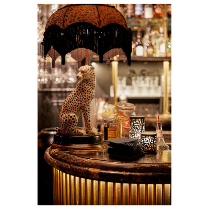 Um bar fracamente iluminado com uma estátua de leopardo no balcão sob uma cúpula de abajur com franjas destaca-se pelo design moderno. Garrafas e guardanapos cercam um elegante castiçal Twine 75 x Ø 75 mm Preto da Duni GmbH, ao lado de uma lanterna de metal, criando uma decoração de mesa convidativa.
.