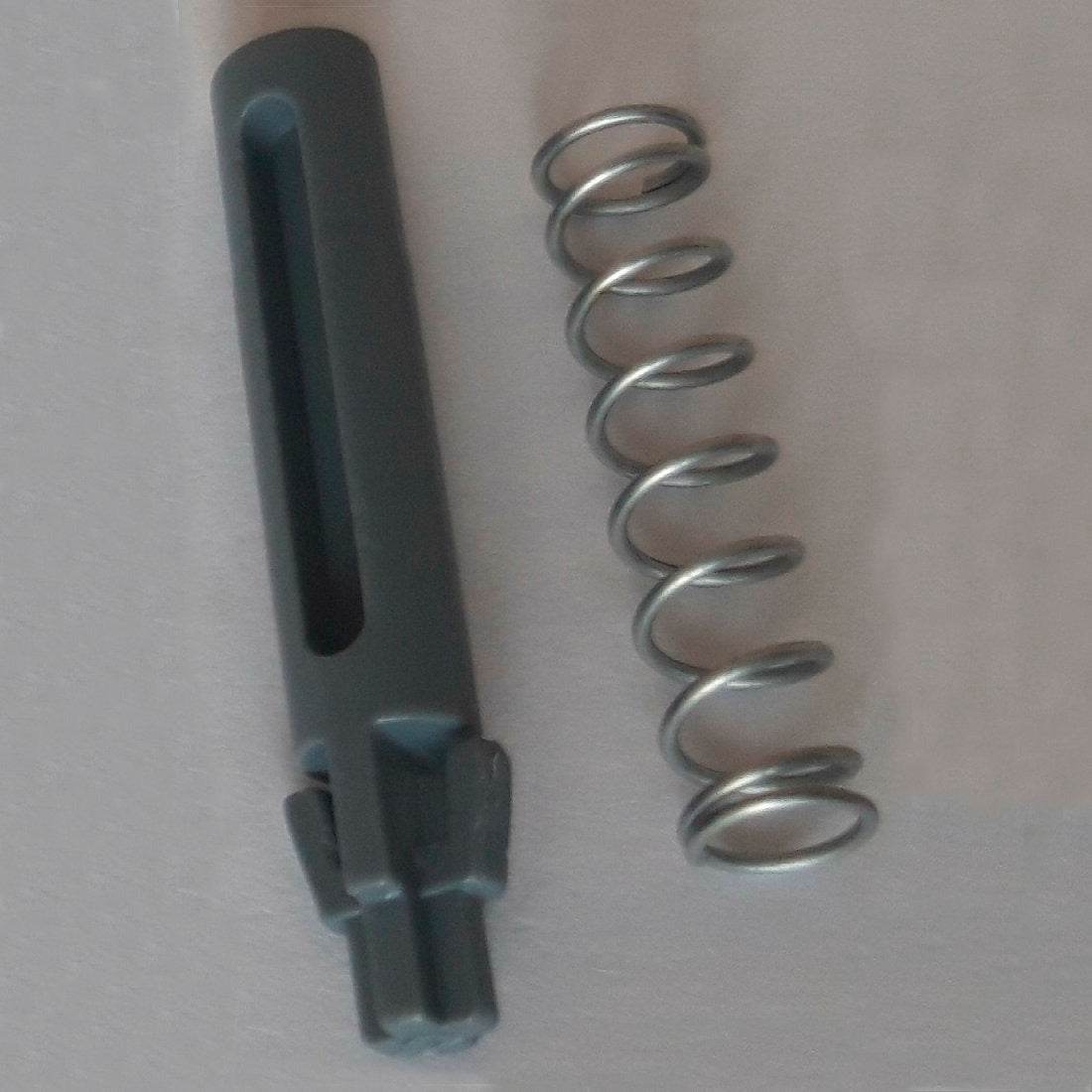 Uma mola de metal e um pino de porta cilíndrico cinza escuro de substituição com ranhuras e entalhes, parte do Pino de Porta & Mola DI Protect (1 peça) da Diversey Deutschland GmbH & Co. OHG, são exibidos sobre uma superfície clara e mostram os materiais robustos para o carrinho DI Protect.