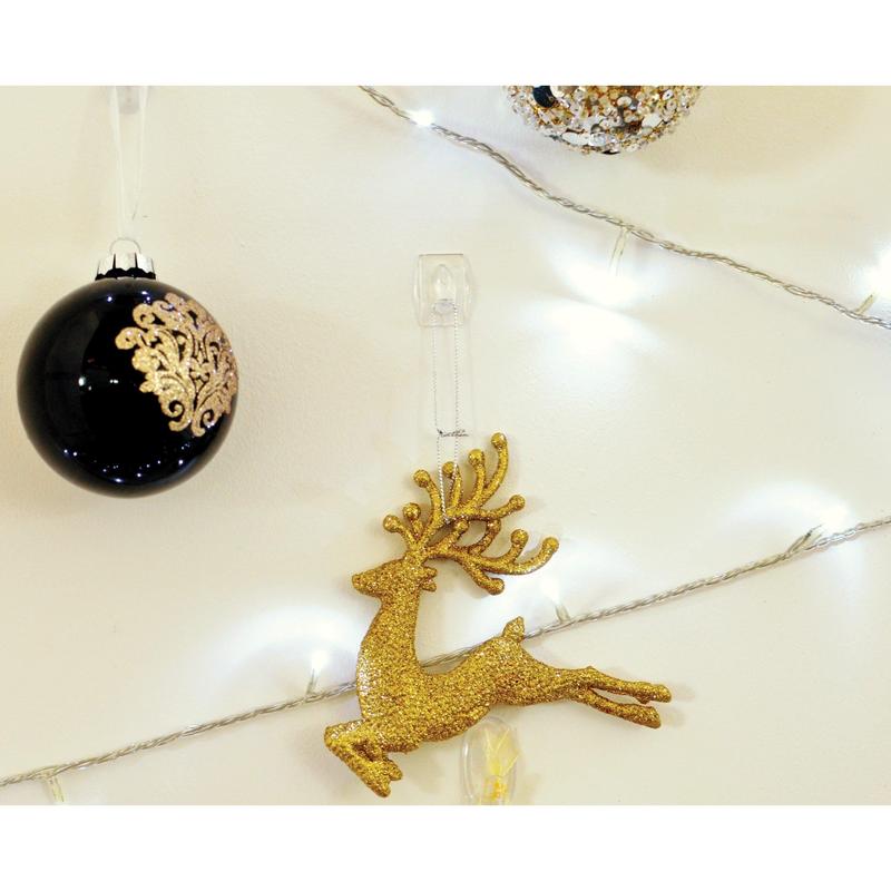 Um ornamento de rena dourada com glitter está pendurado numa parede branca, seguro com um Mini-Gancho Command™ transparente, 17006CLR da 3M Deutschland GmbH, ao lado de um ornamento preto com detalhes dourados e luzes brancas.