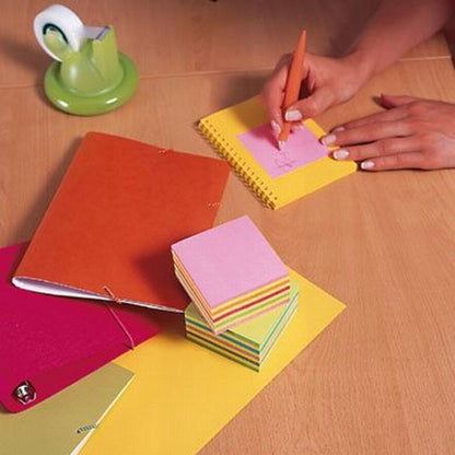 Em uma mesa de madeira, alguém escreve com uma caneta laranja em um bloco de notas Post-it® rosa (76 mm x 76 mm, 450 folhas/bloco, 100% PEFC) da 3M Deutschland GmbH. Perto estão um caderno vermelho, um dispensador de fita adesiva verde, um sino prateado e outros papéis coloridos.