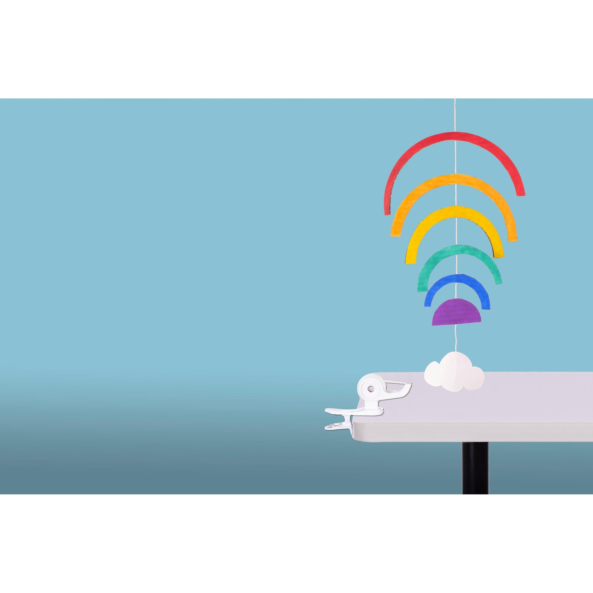 Ein farbenfrohes Papiermobile mit fünf Regenbogenbögen und einer weißen Wolke hängt an der Kante eines weißen Tisches, gesichert durch den 3M Deutschland GmbH Scotch™ Clip & Twist Klebeband-Abroller C19 mit Magic™ Tape, vor einem hellblauen Hintergrund.