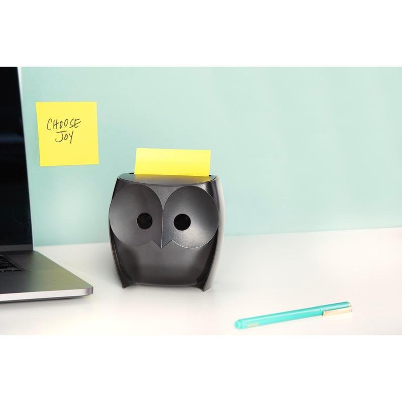 Um dispensador de notas Post-it® Z-Notes preto "coruja" (3M Deutschland GmbH) com notas Super Sticky Z-Notes amarelas está sobre uma mesa branca, ao lado de um laptop e uma caneta azul. Na parede está pendurada uma nota "CHOOSE JOY" sobre um fundo azul claro.