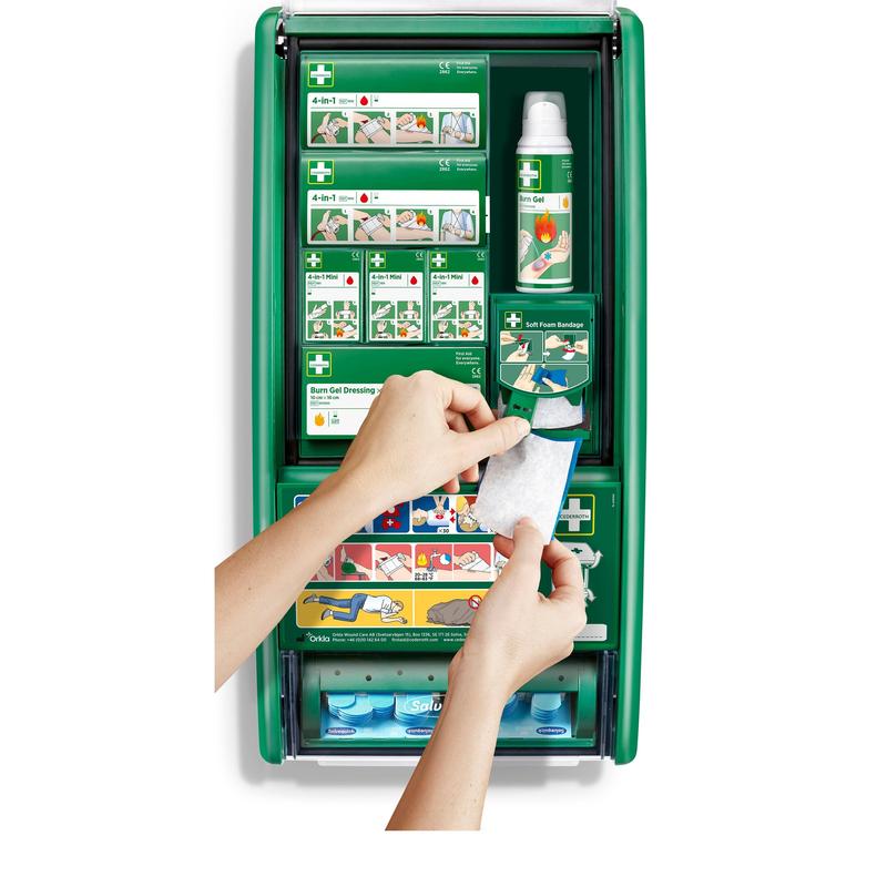 Uma pessoa segura um curativo de gel para queimaduras em frente à estação de primeiros socorros e queimaduras Cederroth (1 embalagem) da Orkla Wound Care AB, uma estação de primeiros socorros verde com material para queimaduras, curativos, frascos e instruções.
