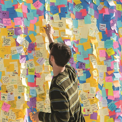 Uma pessoa com camisa listrada está junto a uma parede com mensagens e desenhos manuscritos e pega um cubo de notas adesivas Post-it® da 3M Deutschland GmbH em cores ultra (76x76 mm, 450 folhas, 100% PEFC) para colocá-lo entre os papéis coloridos.