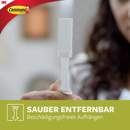 Uma pessoa segura uma Tira de Montagem de Quadros Command™ L 17206, Branco da 3M Deutschland GmbH. Texto: "Removível sem deixar resíduos. Pendurar sem danificar" - ideal para pendurar quadros sem pregos, sobre um fundo verde.