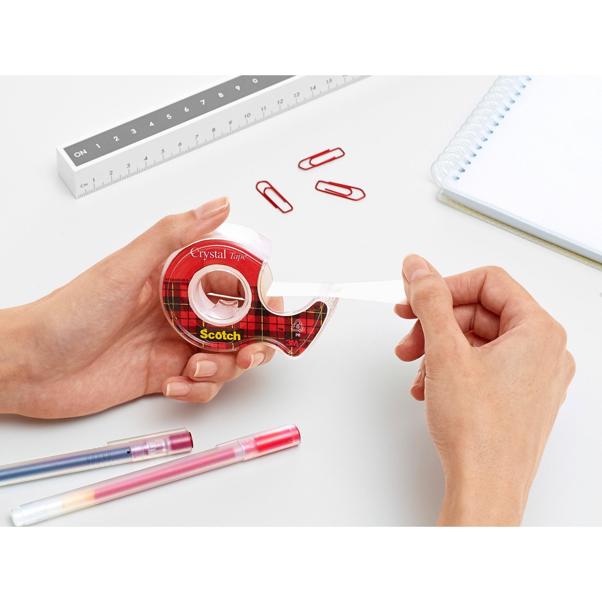 Eine Person zieht Scotch® Crystal Klebeband (19 mm x 7,5 m) aus dem Handabroller der 3M Deutschland GmbH. Auf dem weißen Schreibtisch liegen rote Büroklammern, ein Lineal, ein Notizbuch und zwei farbige Stifte.