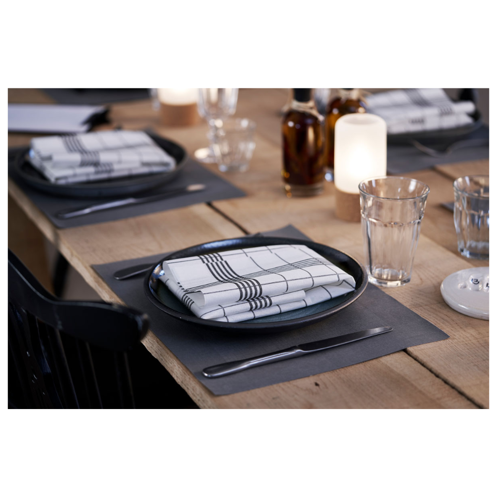 Um conjunto de mesa de jantar é composto por pratos em Linnea Black da Duni GmbH e guardanapos xadrez. Ao fundo, três garrafas de vidro estão ao lado de velas acesas. Copos de água e talheres são elegantemente apresentados sobre um conjunto de mesa Duni Dunicel® 30 x 40 cm, destacando a simplicidade elegante da mesa de madeira clara.