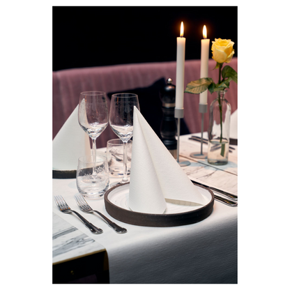 Uma mesa de jantar está elegantemente posta com dois guardanapos Duni Dunilin® 48 x 48 cm Branco, dobrados em triângulos sobre os pratos. Duas grandes velas acesas e uma rosa amarela em um vaso de vidro adornam o fundo. Taças de vinho, um copo de água e talheres completam este ambiente sofisticado com charme semelhante a tecido.