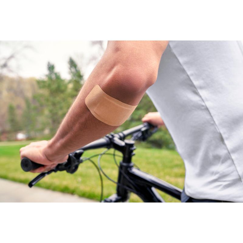 Uma pessoa vestindo uma camisa branca e a andar de bicicleta ao ar livre tem um penso Nexcare™ Ultra Stretch MAXI Comfort Flexible (50 mm x 101 mm) da 3M Deutschland GmbH no braço nu; ao fundo vê-se relva verde e árvores.