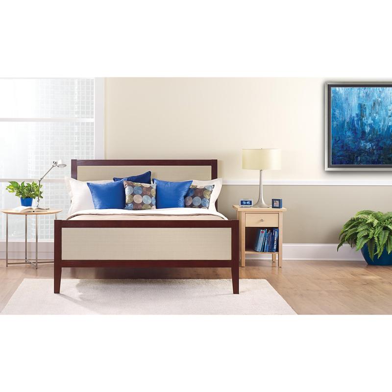 Uma cama bem feita com almofadas brancas e azuis está em frente a uma parede clara, que está coberta com a fita adesiva para pintura ScotchBlue™ "Multi-Surface" da 3M Deutschland GmbH. Sobre uma mesa de cabeceira, há uma lâmpada, um relógio, livros e uma planta em vaso ao lado da janela clara.