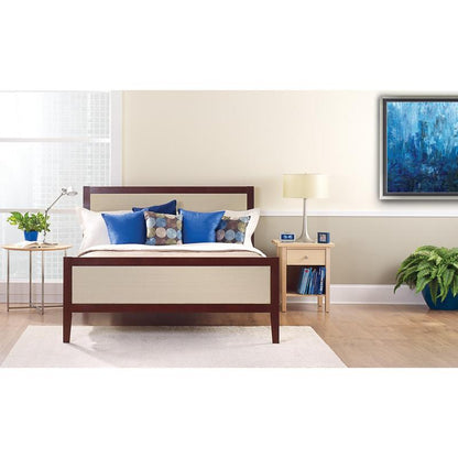 Uma cama bem feita com almofadas brancas e azuis está em frente a uma parede clara, que está coberta com a fita adesiva para pintura ScotchBlue™ "Multi-Surface" da 3M Deutschland GmbH. Sobre uma mesa de cabeceira, há uma lâmpada, um relógio, livros e uma planta em vaso ao lado da janela clara.