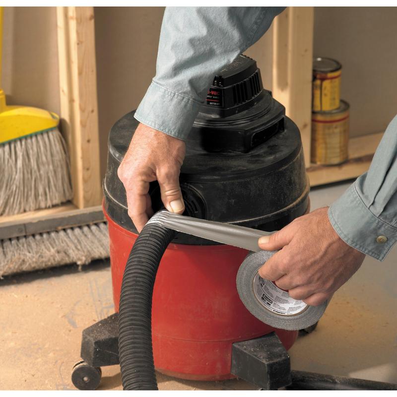 Uma pessoa envolve a fita adesiva universal Scotch® 2904 (10 m x 48 mm) da 3M Deutschland GmbH em torno da conexão da mangueira de um aspirador de pó úmido/seco vermelho e preto, ao fundo vê-se uma vassoura e latas de tinta.