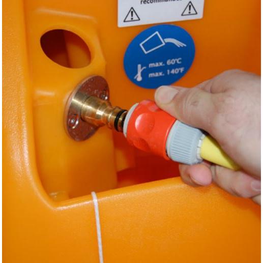 Uma mão conecta uma mangueira a uma peça de conexão vermelha e cinza com um encaixe de metal em um TASKI swingo 2100 ?icro AquaStop laranja da Diversey Deutschland GmbH & Co. OHG. Um adesivo azul próximo indica uma temperatura máxima de 60°C/140°F.