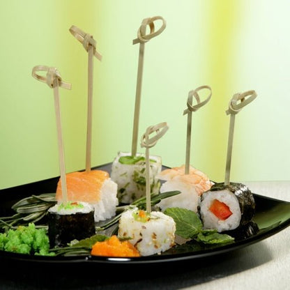 Um prato preto com vários sushis, cada peça espetada com um PAPSTAR 40 espeto para fingerfood "pure" da PAPSTAR GmbH, decorado com ervas e ovas, sobre uma superfície verde-amarela – ideal para destacar acessórios de catering.