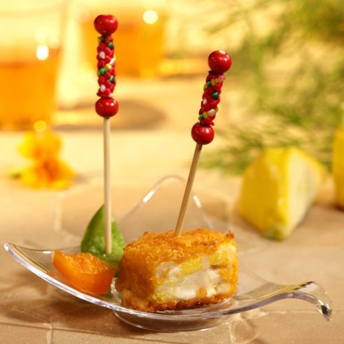 Em um prato transparente em forma de folha, há um cubo de alimentos empanados e fritos com um PAPSTAR 250 palito de finger food da PAPSTAR GmbH, além de uma fatia de laranja e uma fatia verde. Antes do fundo levemente desfocado, pode-se ver uma fatia de limão e verduras.