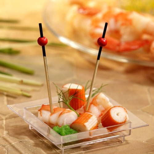 Em uma tigela quadrada e transparente, há imitação de caranguejo, camarões, uma guarnição verde e dois PAPSTAR 250 palitos de finger food da PAPSTAR GmbH com pérolas vermelhas. Ao fundo, podem-se ver camarões desfocados em um prato.
