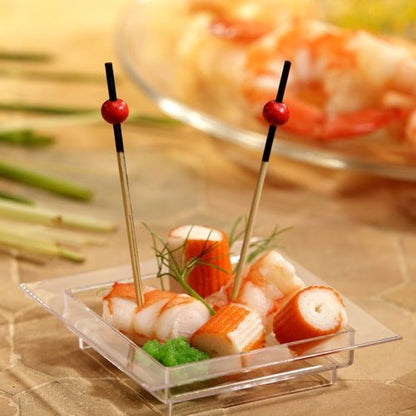Em uma tigela quadrada e transparente, há imitação de caranguejo, camarões, uma guarnição verde e dois PAPSTAR 250 palitos de finger food da PAPSTAR GmbH com pérolas vermelhas. Ao fundo, podem-se ver camarões desfocados em um prato.