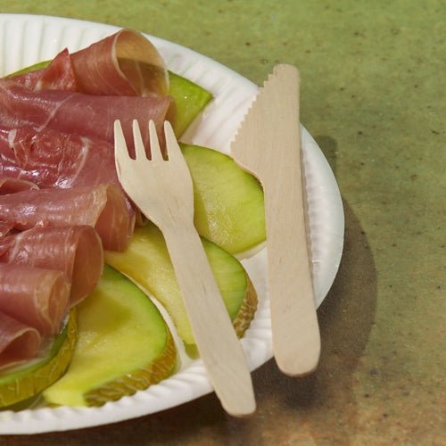 Um prato de papel branco com fatias de maçã verde e carne curada enrolada está sobre uma superfície texturizada em verde, ao lado um garfo de madeira e uma faca de madeira PAPSTAR "pure" 16,5 cm da PAPSTAR GmbH, ecológica e estável.