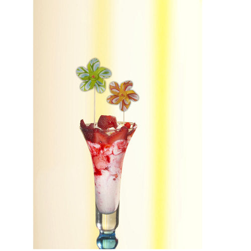 Um copo alto contém uma sobremesa rosa e branca, coberta com calda de morango e um PAPSTAR palito decorativo "Cata-vento" (17,5 cm, cores sortidas), que enriquece a decoração da festa de forma lúdica. O conjunto da PAPSTAR GmbH contém 50 palitos em uma bandeja de papelão.