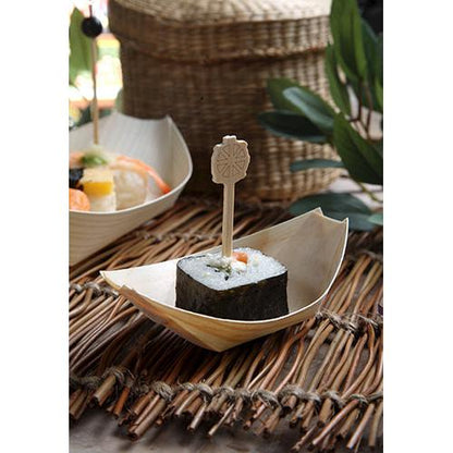 Um pedaço de sushi, envolto em alga marinha e preso com um espeto de madeira, é servido em um barquinho de madeira "pure" da PAPSTAR 50 bandejas para finger food da PAPSTAR GmbH, apresentado sobre uma esteira de palha com folhas e cestos de vime ao fundo.