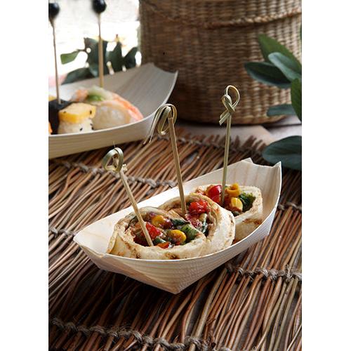 Três rolinhos de pão sírio recheados com vegetais em espetos são servidos como finger food em uma bandeja de madeira "barquinho" da PAPSTAR 50 da PAPSTAR GmbH sobre um jogo americano trançado. Ao fundo, um outro prato está desfocado.