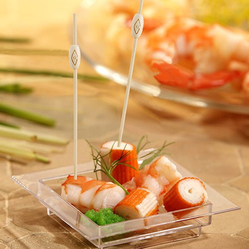 Numa bandeja quadrada de plástico com imitação de caranguejo, camarões, ovas verdes e decoração, estão representados dois PAPSTAR 40 espetos para fingerfood "pure" da PAPSTAR GmbH, ao fundo vê-se desfocado um prato semelhante de frutos do mar.