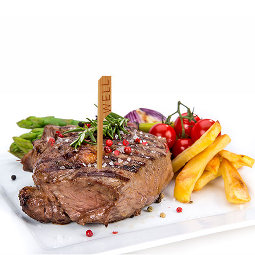 Um bife grelhado com a inscrição "WELL" e um PAPSTAR 250 marcador de bife (9 cm, PAPSTAR GmbH) é servido num prato branco com espargos, tomates cereja, cebolas fritas e batatas fritas, guarnecido com alecrim e grãos de pimenta.