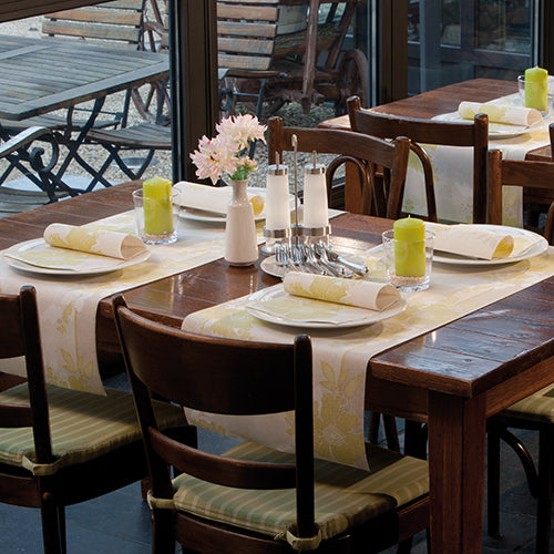 Ein hölzerner Esstisch für vier Personen mit weißen Tellern, PAPSTAR 50 Servietten "ROYAL Collection" 1/4-Falz 40 x 40 cm grün von PAPSTAR GmbH, grünen Kerzen, Glaswaren, Salz- und Pfefferstreuern und einer Vase mit rosa Blumen am Fenster.