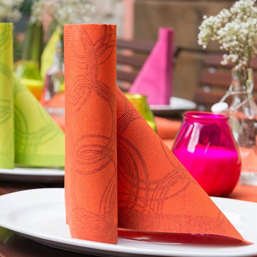 Um guardanapo laranja PAPSTAR 50 guardanapos "ROYAL Collection" 1/4-dobra 40 x 40 cm nectarina está em pé sobre um prato branco, com decoração colorida ROYAL Collection e flores ao fundo desfocadas.