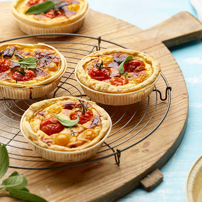 Três pequenas quiches de vegetais com tomate cereja e ervas estão em um suporte redondo de arame sobre uma tábua de madeira, rodeadas por folhas de ervas frescas e PAPSTAR 50 formas de papel "puras" castanhas da PAPSTAR GmbH.