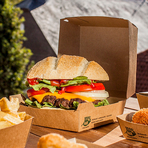 Um cheeseburger com alface, tomate, cebola e queijo cheddar está ao ar livre em uma caixa de hambúrguer PAPSTAR GmbH feita de papelão (9x18,5x18,5 cm, "100% Justo", 25 unidades). Batatas fritas e outros acompanhamentos são servidos separadamente ao lado.
