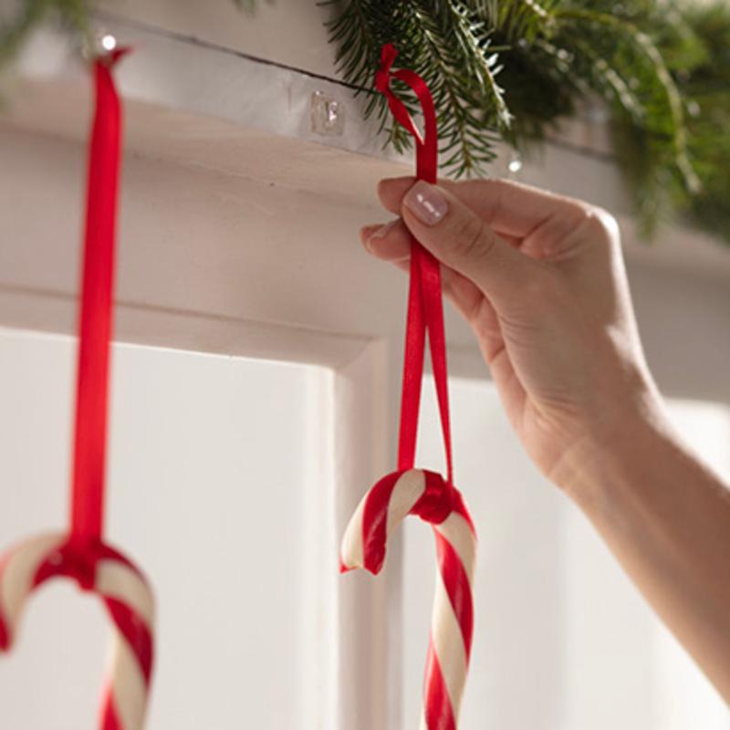 Uma mão pendura um bastão de açúcar com uma fita vermelha num Mini-Gancho Command™ transparente, com tiras transparentes (3M Deutschland GmbH), que está fixado sob uma prateleira decorada com ramos verdes. Outro bastão de açúcar já está pendurado no gancho ao lado.