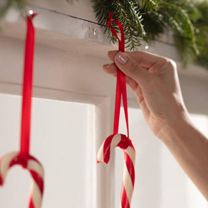 Uma mão pendura um bastão de açúcar com uma fita vermelha num Mini-Gancho Command™ transparente, com tiras transparentes (3M Deutschland GmbH), que está fixado sob uma prateleira decorada com ramos verdes. Outro bastão de açúcar já está pendurado no gancho ao lado.