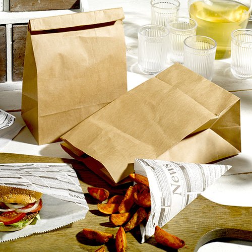 Dois PAPSTAR 250 sacos de fundo quadrado de papel kraft marrom da PAPSTAR GmbH, um saco de papel com design de jornal com batatas fritas e um hambúrguer parcialmente embrulhado em papel de jornal estão com quatro copos e uma jarra sobre uma mesa clara.