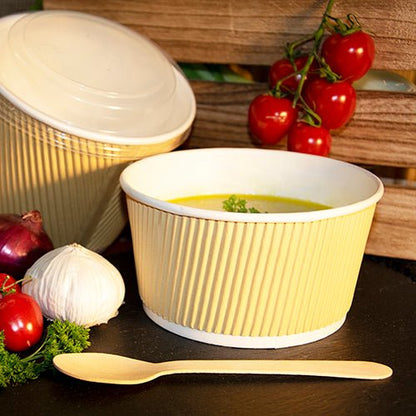 Um copo de sopa redondo castanho/branco PAPSTAR, 11 cm Ripple Wall (25 peças) da PAPSTAR GmbH está cheio de sopa amarela, ao lado uma colher de madeira, alho, cebola, tomates cereja no ramo, salsa e outra tigela fechada ao fundo.