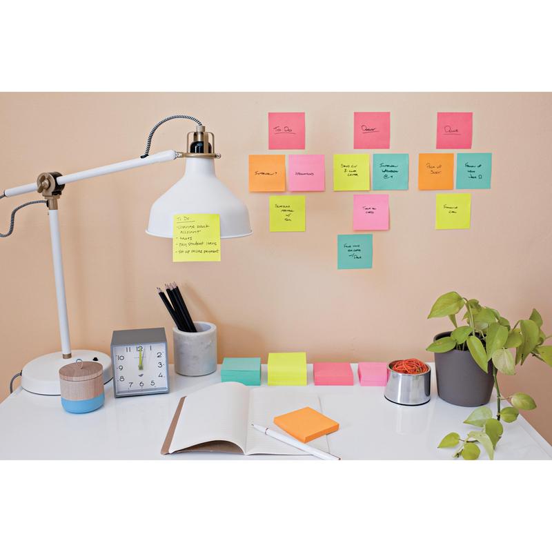 Uma mesa organizada com lâmpada, relógio, planta, porta-canetas, um caderno aberto e canetas. Na parede acima da mesa estão fixados cubos de notas adesivas Post-it®, cores ultra, da 3M Deutschland GmbH, para lembretes e tarefas manuscritas.