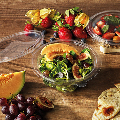 Uma tigela de delicatessen e saladas Starpak 50 com tampa dobrável, R-PET transparente e redonda, contém uma salada de melão e figo sobre uma mesa de madeira com morangos, uvas, pão, cerejas e frutas. Outro recipiente Starpak R-PET com frutas está próximo.