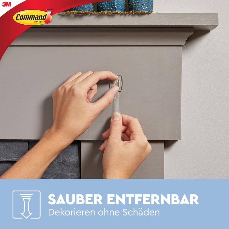 Uma pessoa puxa uma tira adesiva transparente de uma lareira cinza e mostra como os Mini-Ganchos Command™ transparentes, com tiras transparentes (17006CLR) da 3M Deutschland GmbH decoram e fixam sem deixar marcas. Os logos da Command e 3M são visíveis.