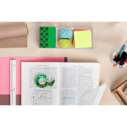 Um espaço de trabalho com um livro aberto, papel milimetrado, bolas de fio colorido, notas adesivas, canetas em um copo, borrachas, um rolo de papel pardo e um rolo de Fita Invisível Scotch® Magic™ da 3M Deutschland GmbH sobre uma mesa de madeira clara.