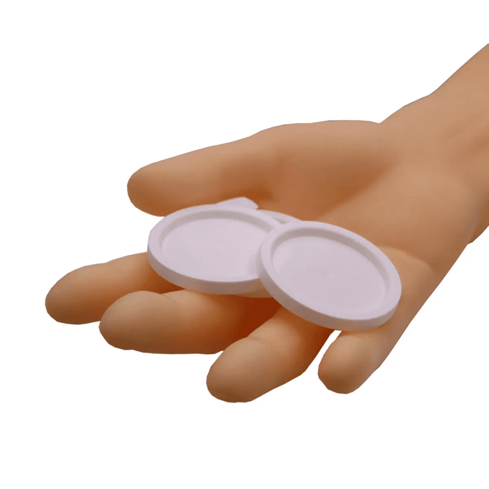 Uma mão segura duas tampas redondas AMPri para copos de medicamentos, objetos brancos sobre um fundo branco simples.
