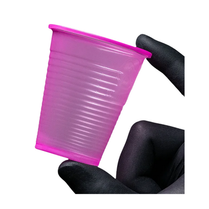 Um pequeno copo rosa AMPri Dental PP Mundspülbecher de polipropileno é segurado entre dois dedos enluvados diante de um fundo branco. Este copo descartável de enxaguante bucal da AMPri Handelsgesellschaft mbH, frequentemente encontrado em consultórios dentários, possui uma superfície texturizada com ranhuras horizontais.