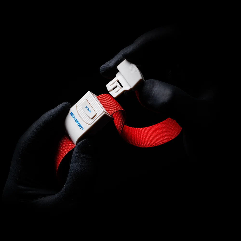 Mãos enluvadas, que lembram um torniquete AMPri MED-COMFORT, seguram a fivela de um cinto de segurança vermelho brilhante diante de um fundo sombreado. Na fivela, estão visíveis a palavra "Blue" e a letra "B".