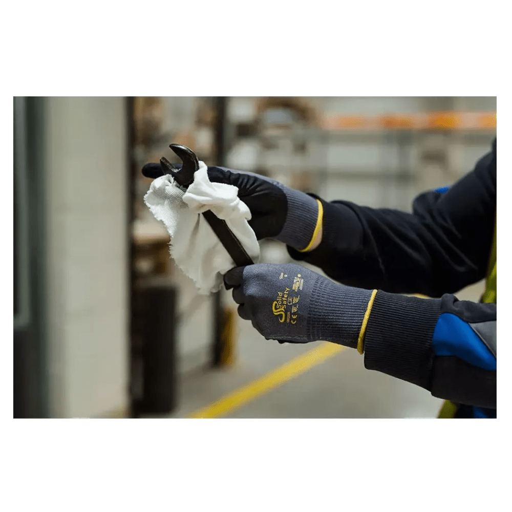 Uma pessoa com luvas de montagem ultraleves AMPri SolidSafety Tough Air da AMPri Handelsgesellschaft mbH limpa cuidadosamente uma chave inglesa com um pano branco em ambientes internos. As luvas cinzentas, conhecidas pelo seu nível de resistência à abrasão 4, destacam-se das prateleiras e equipamentos industriais desfocados ao fundo.
