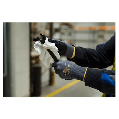 Uma pessoa com luvas de montagem ultraleves AMPri SolidSafety Tough Air da AMPri Handelsgesellschaft mbH limpa cuidadosamente uma chave inglesa com um pano branco em ambientes internos. As luvas cinzentas, conhecidas pelo seu nível de resistência à abrasão 4, destacam-se das prateleiras e equipamentos industriais desfocados ao fundo.