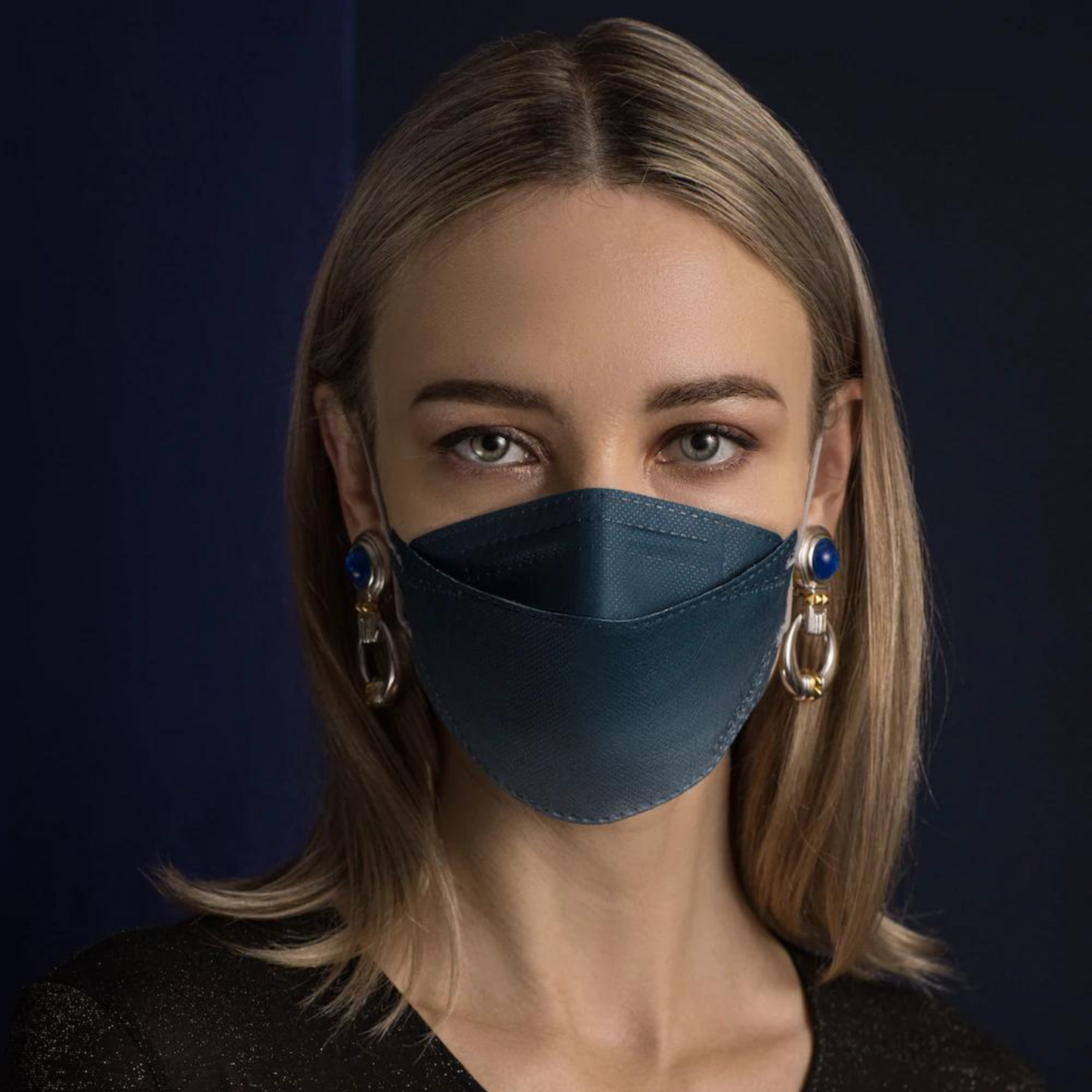 Uma pessoa com cabelo longo usa uma máscara facial de designer Altruan com filtro de partículas em azul, que oferece proteção eficaz de acordo com o padrão FFP2. Ela combina com brincos decorativos grandes em frente a um fundo escuro e usa uma camisa preta.