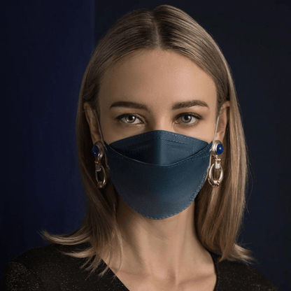 Uma pessoa com cabelo longo usa uma máscara facial de designer Altruan com filtro de partículas em azul, que oferece proteção eficaz de acordo com o padrão FFP2. Ela combina com brincos decorativos grandes em frente a um fundo escuro e usa uma camisa preta.