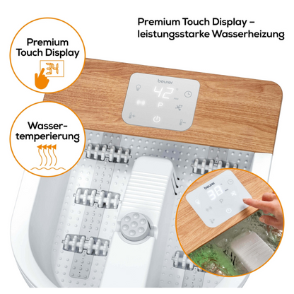 Um spa para pés da marca Beurer FB 65 Wellness Spa para Pés da Beurer GmbH tem uma base branca e uma placa de madeira, com um display digital touch que mostra uma temperatura de 42 °C. As etiquetas dizem "Aquecimento de água" e "Display Touch Premium". Uma mão ajusta o display e rolos de massagem para massagem vibratória e de bolhas são visíveis.